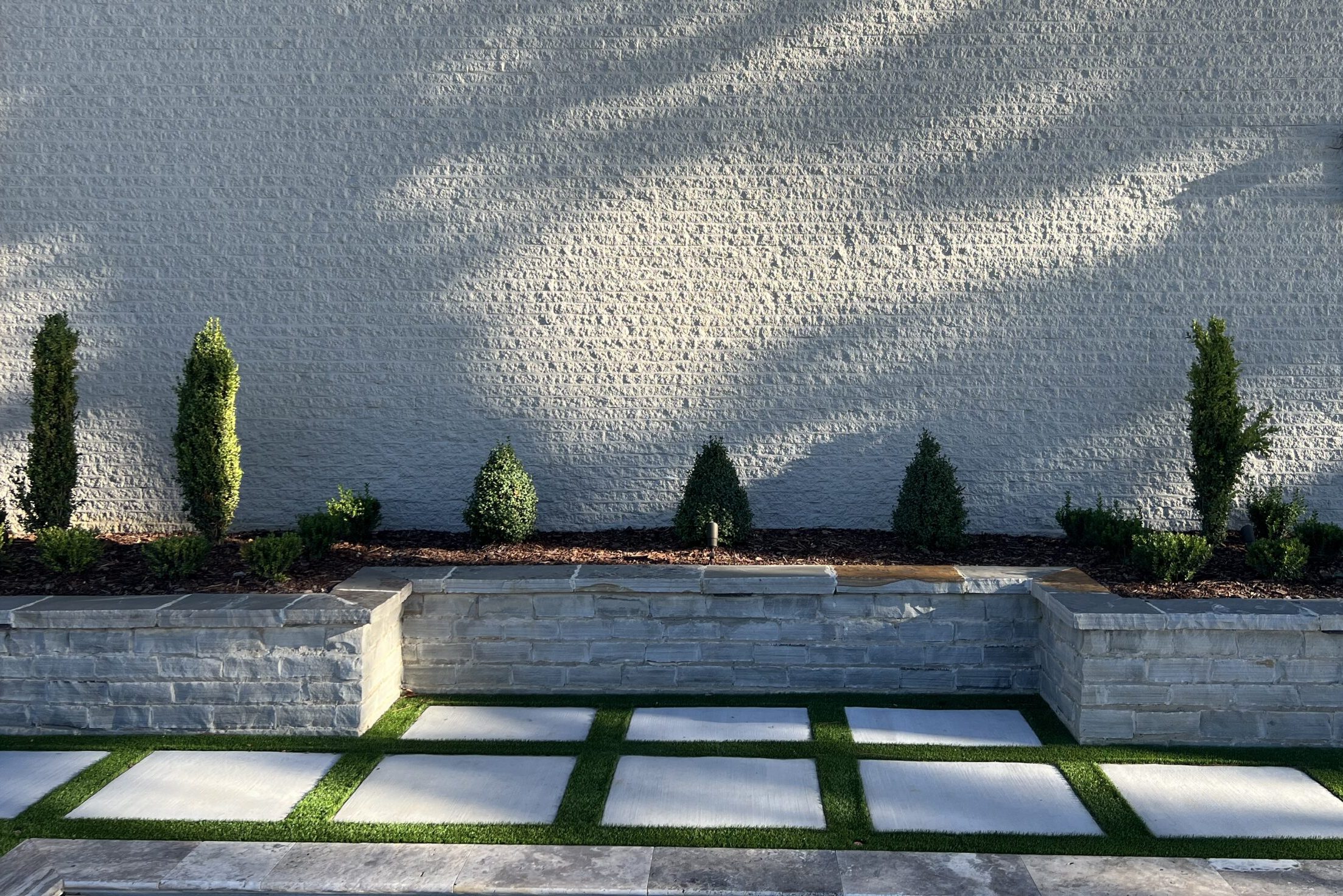 Modern backyard patio featuring a stone retaining wall planter, artificial turf strips between concrete pavers, and manicured shrubs against a gray brick wall.