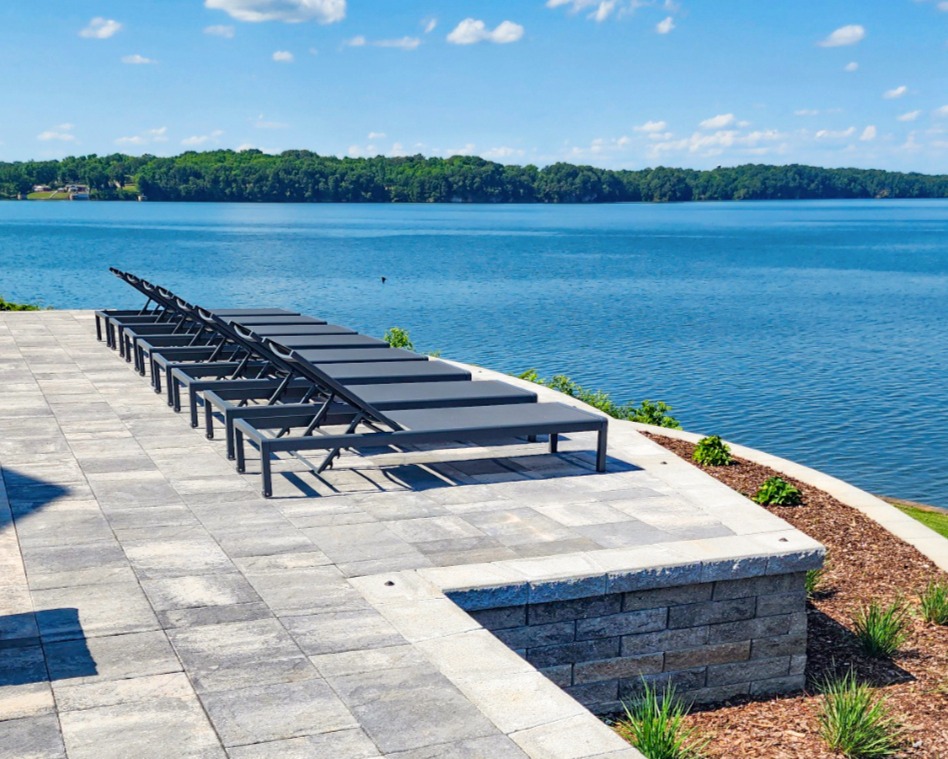 Custom paver patio and engineered retaining wall overlooking a river, with modern lounge chairs.